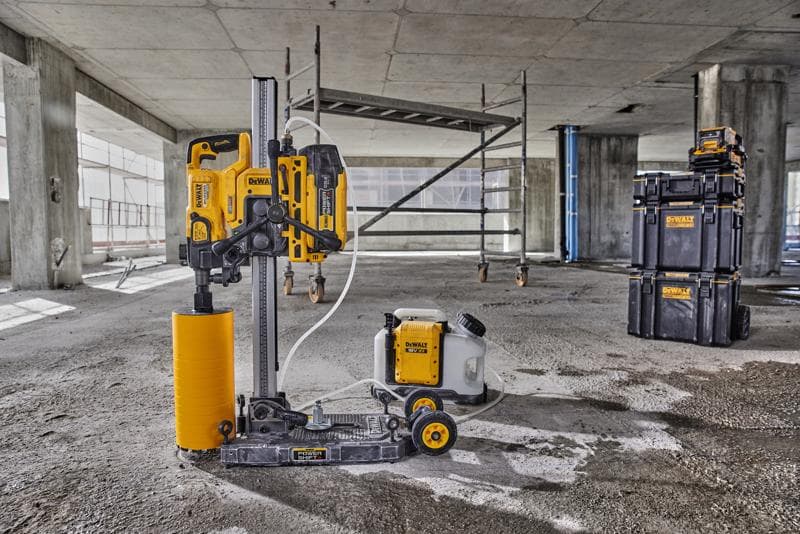Una máquina de perforación de núcleo DEWALT con un accesorio de broca cilíndrica amarilla y un depósito de agua conectado está colocada en el suelo de una obra de construcción de cemento. Al fondo se ven andamios y cajas apilables de DEWALT.