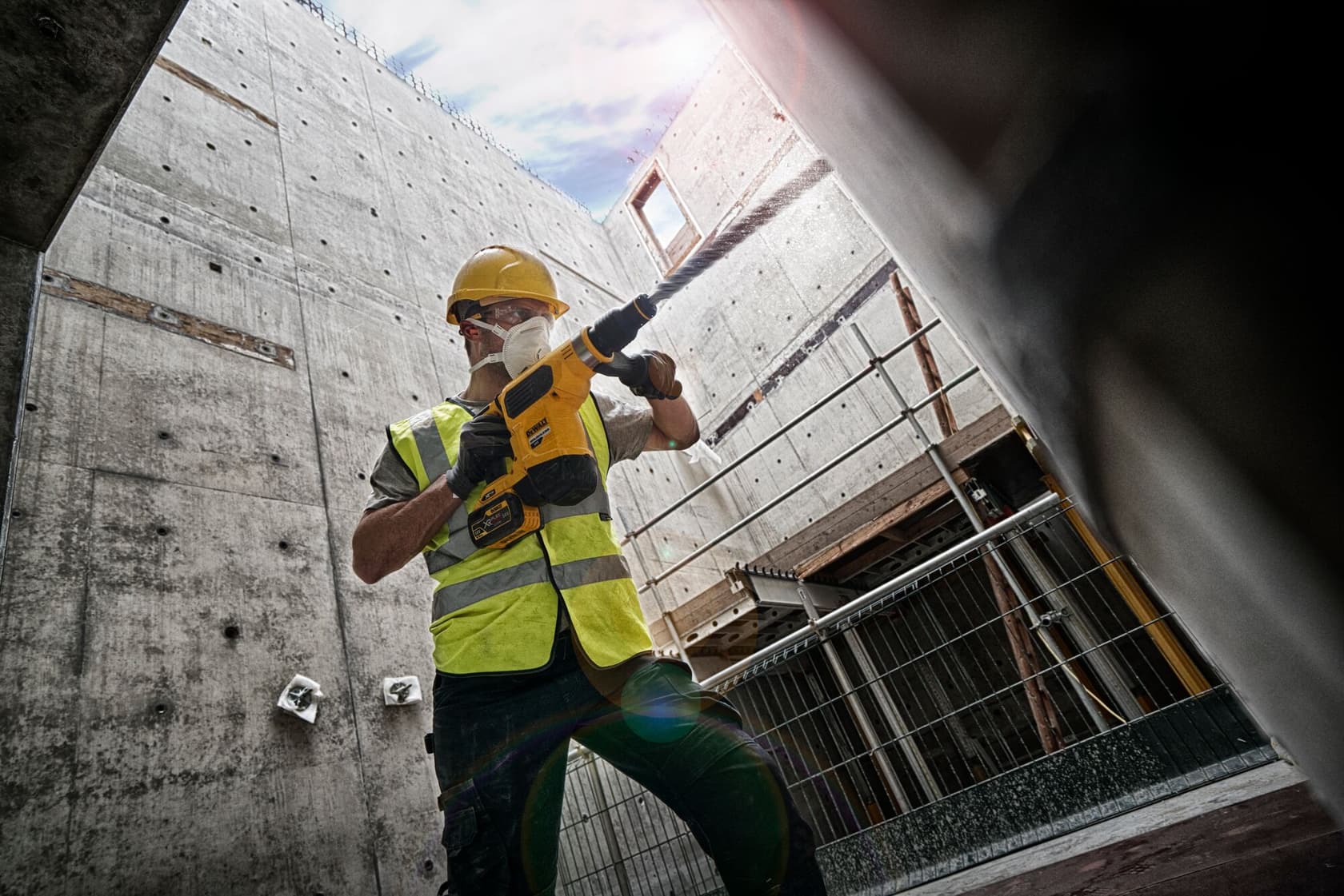 Worker using a DEWALT rotary hammer drill on a construction site, wearing helmet, vest, and mask.