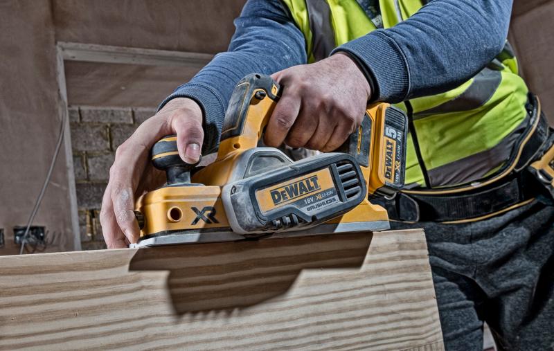 Persona usando un cepillo inalámbrico DEWALT DCP580N sobre madera en un sitio de construcción.
