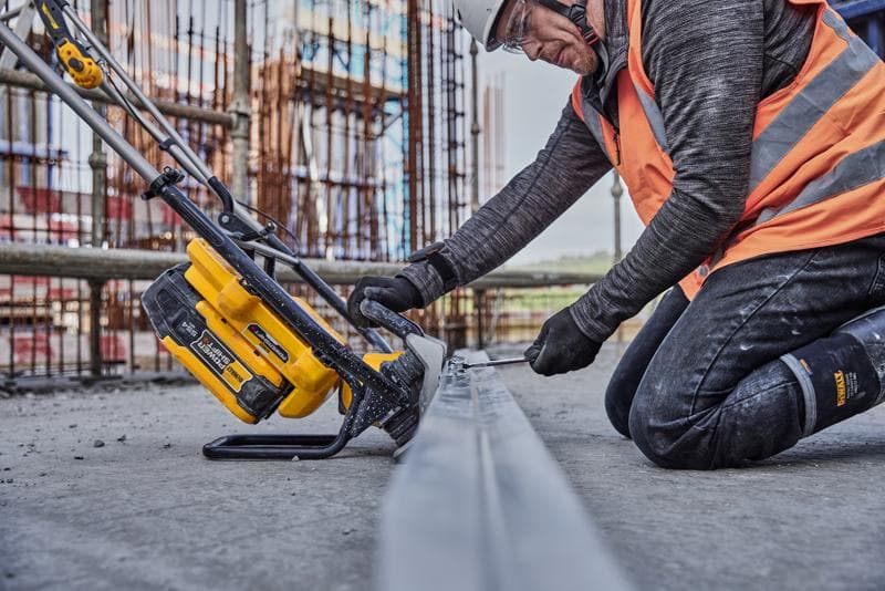 Trabajador usa herramienta DEWALT amarilla para cortar metal en obra, lleva chaleco naranja y equipo de seguridad.
