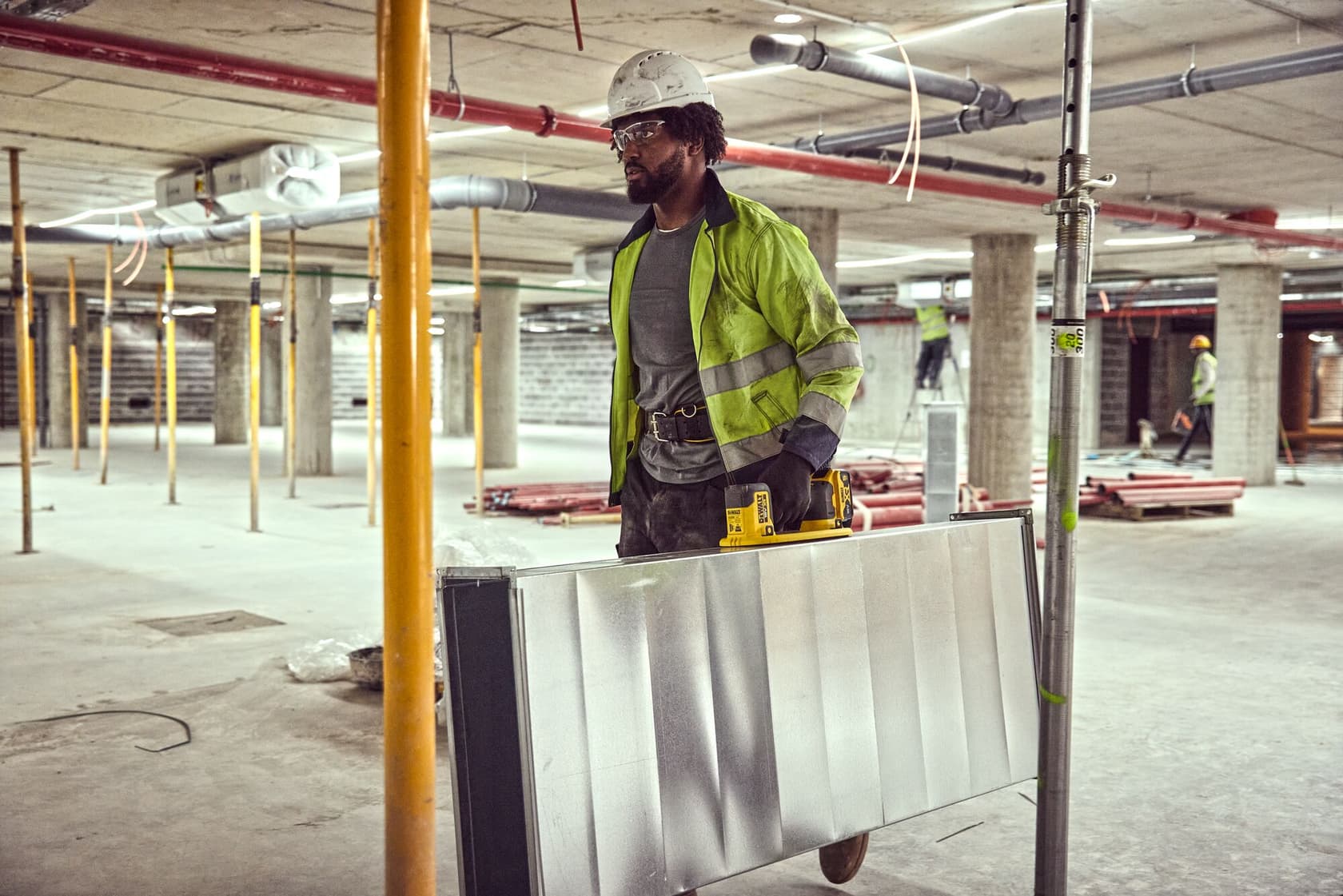 Trabajador con chaqueta reflectante y casco lleva placa metálica y herramienta DEWALT en obra.