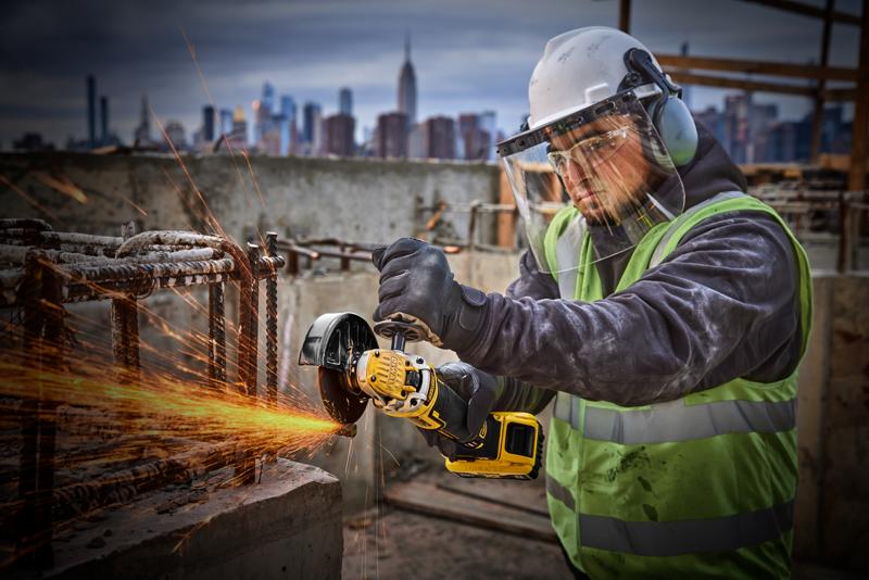 Trabajador usa amoladora DEWALT DCG405P2 en obra, con equipo de seguridad, ciudad al fondo.