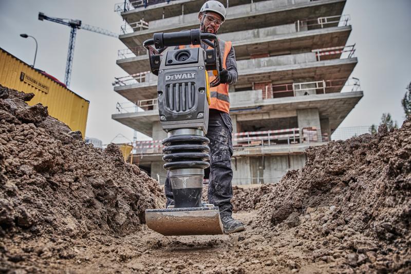 Compactador DEWALT en obra, usado por un trabajador con equipo de seguridad, rodeado de tierra y edificios.