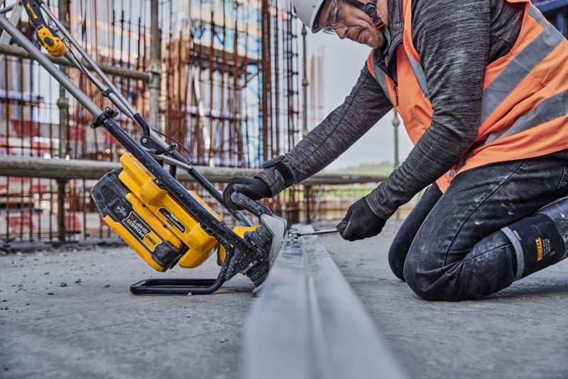 Trabajador usa herramienta DEWALT amarilla para cortar metal en obra, lleva chaleco naranja y equipo de seguridad.
