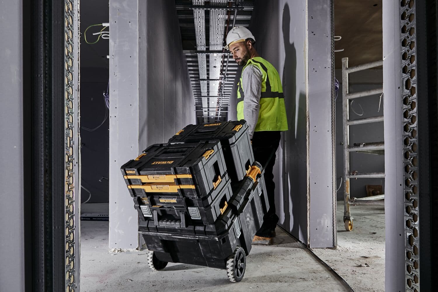 Trabajador lleva cajas de herramientas DEWALT apiladas con ruedas por un pasillo estrecho en obra.