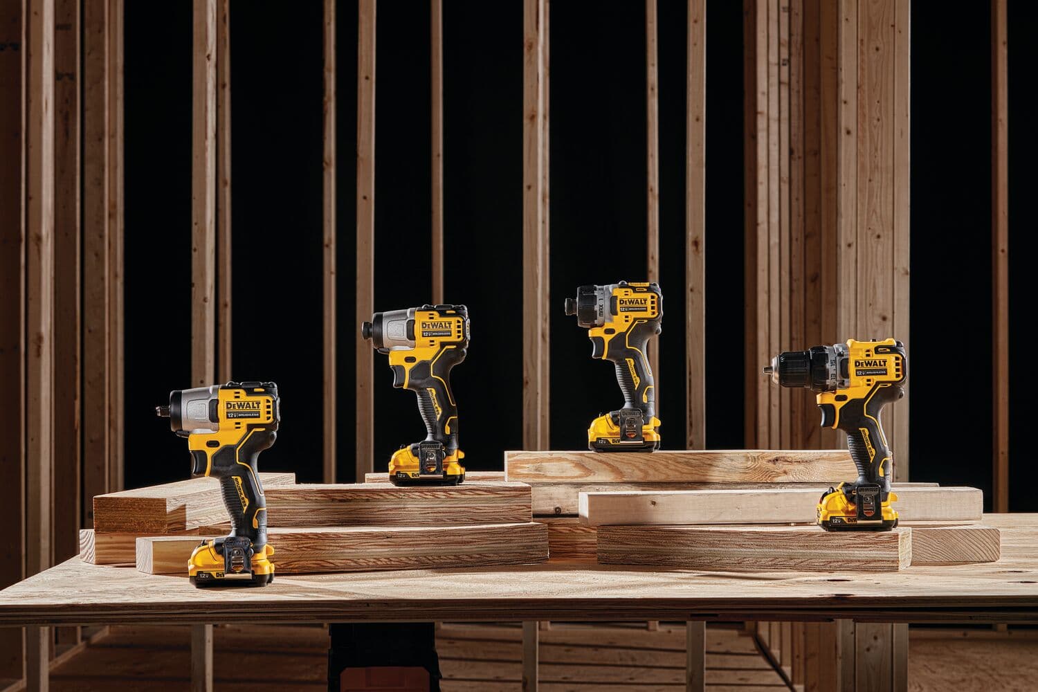 Cuatro taladros inalámbricos DEWALT están exhibidos sobre tablones de madera en un entorno de construcción con estructura de madera en el fondo.