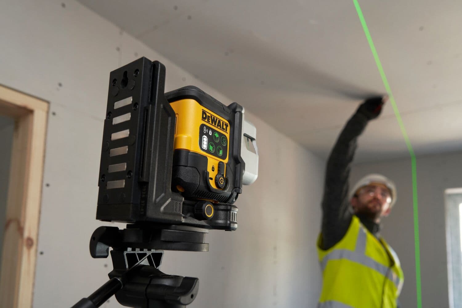 Nivel láser DEWALT proyecta línea verde, persona con chaleco trabaja en techo de sala sin terminar.