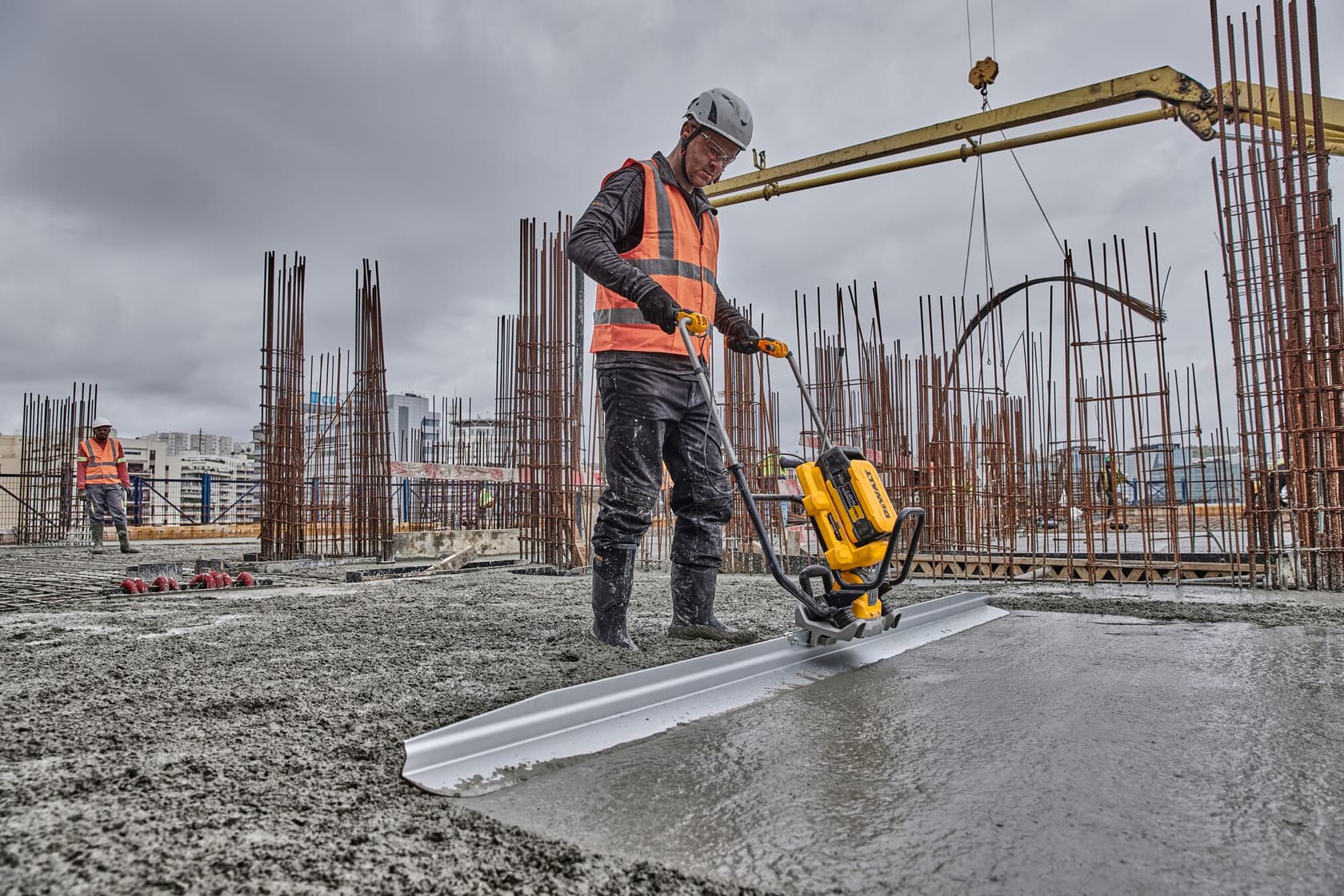 Trabajador usa DEWALT para alisar concreto fresco en obra, con varillas de acero y maquinaria visibles.