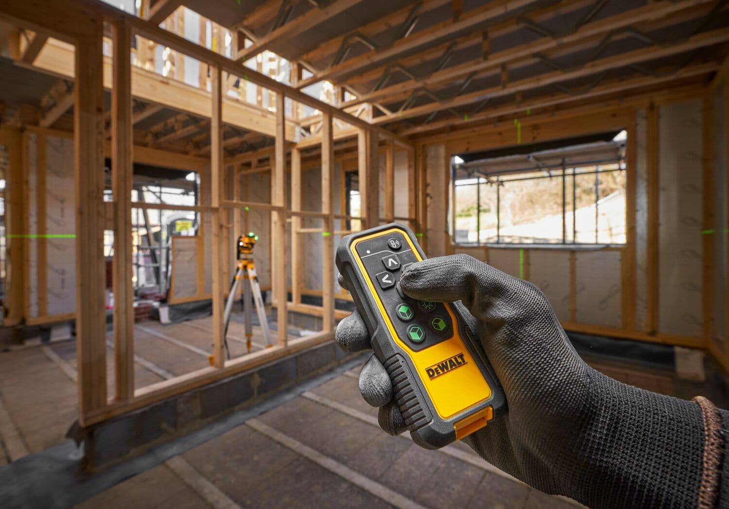 Mano con guante sostiene control DEWALT en obra de madera. Nivel láser visible en el fondo.