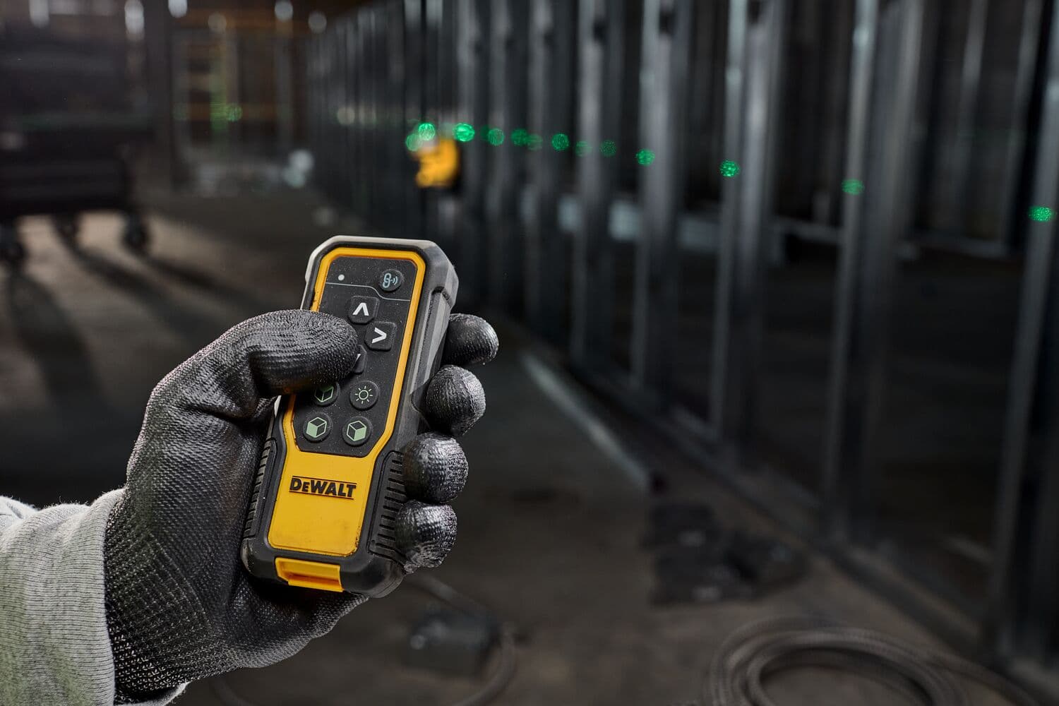 Mano con guante sostiene control remoto DEWALT láser, puntos verdes visibles en obra.