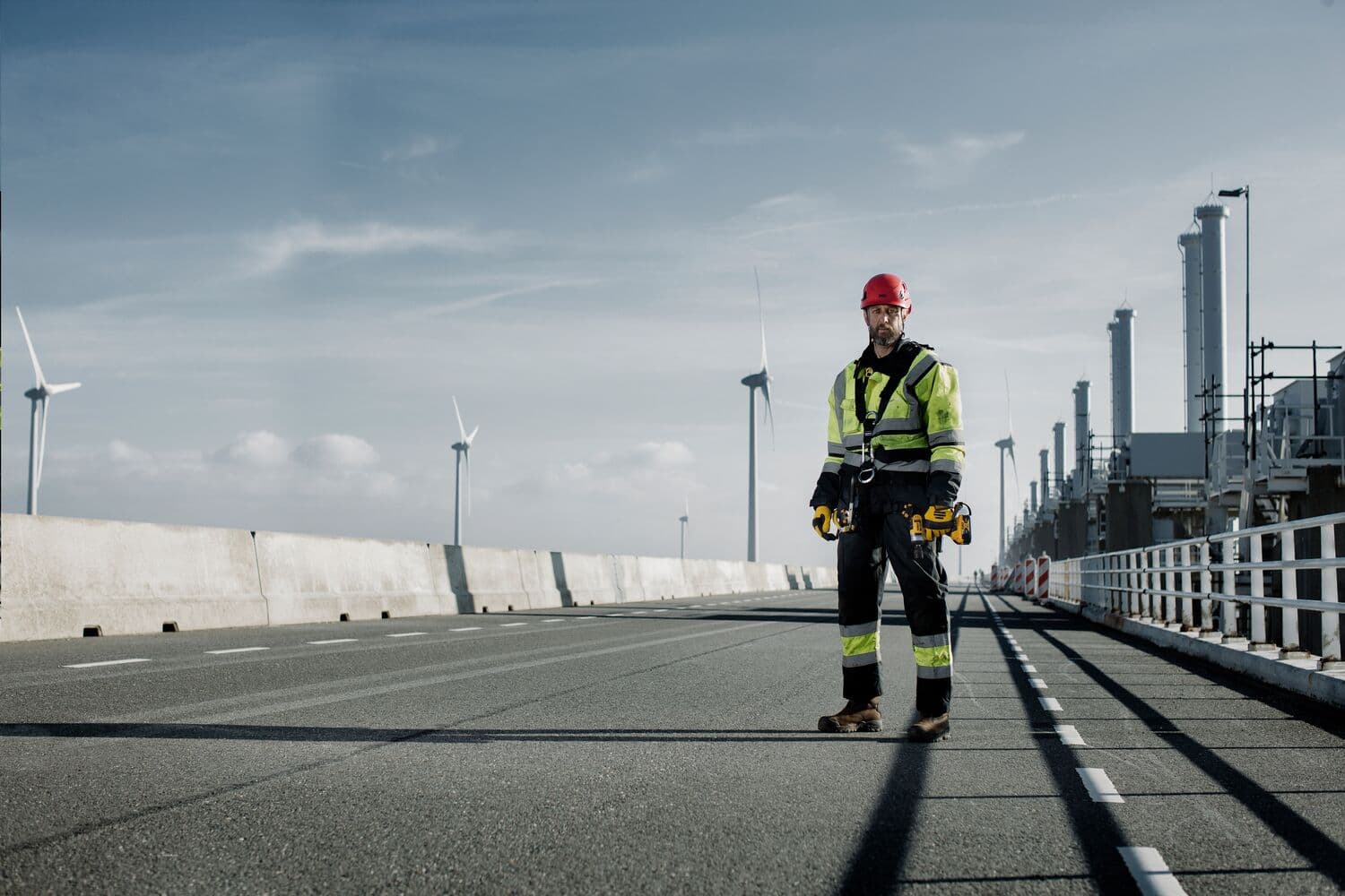 Trabajador con equipo de seguridad en carretera industrial, sostiene herramientas. Fábricas y turbinas de fondo.