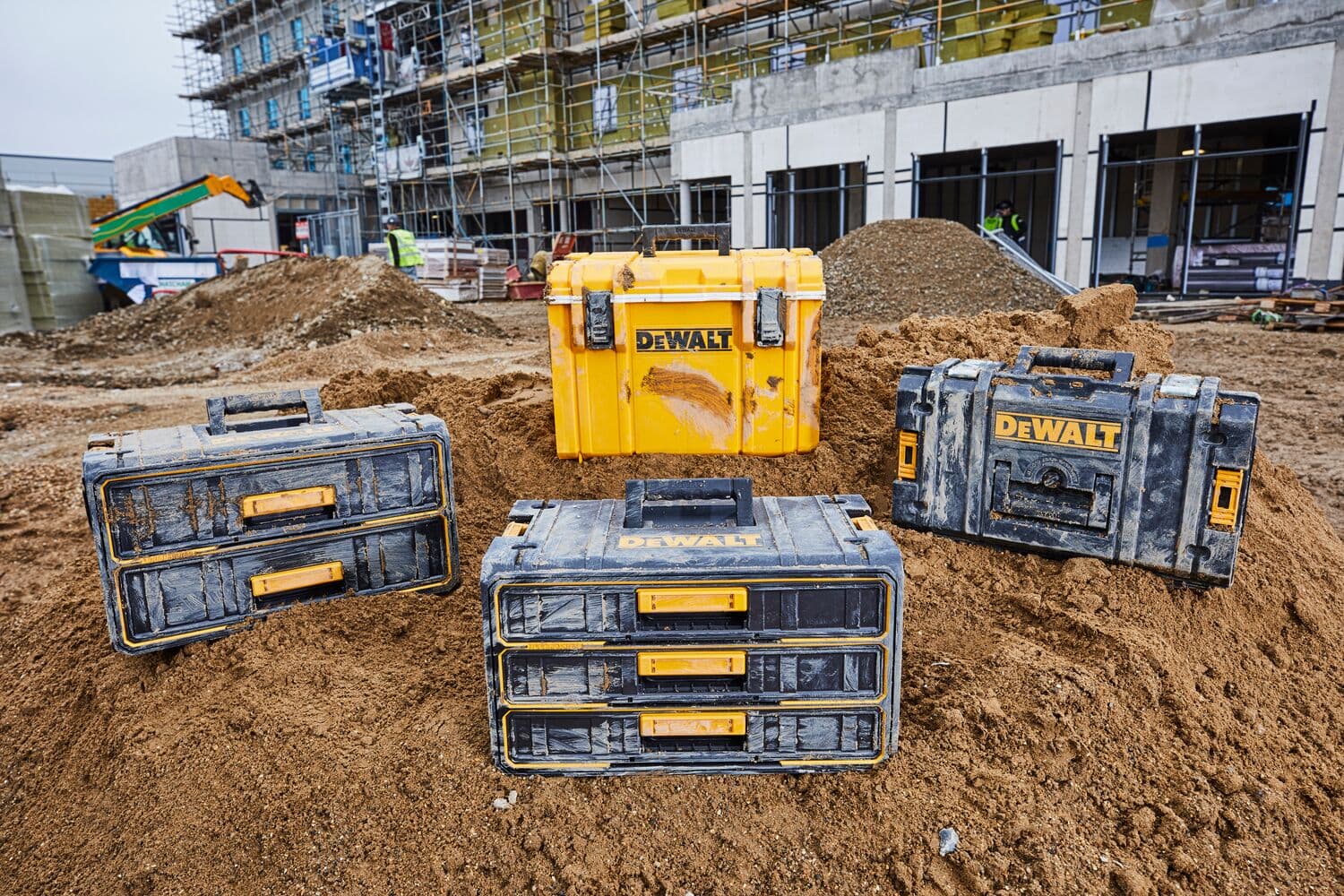 Cuatro cajas de herramientas DEWALT sobre tierra en una obra, con edificio y trabajadores al fondo.
